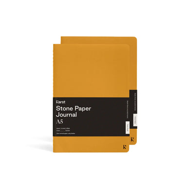 Two turmeric yellow journals stagger stacked on top of each other; Front journal has large black rectangular paper wrap half way down reads 